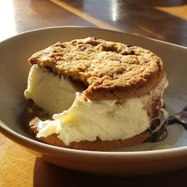 A partially eaten ice cream sandwich with chocolate chip cookies and vanilla ice cream in a bowl.