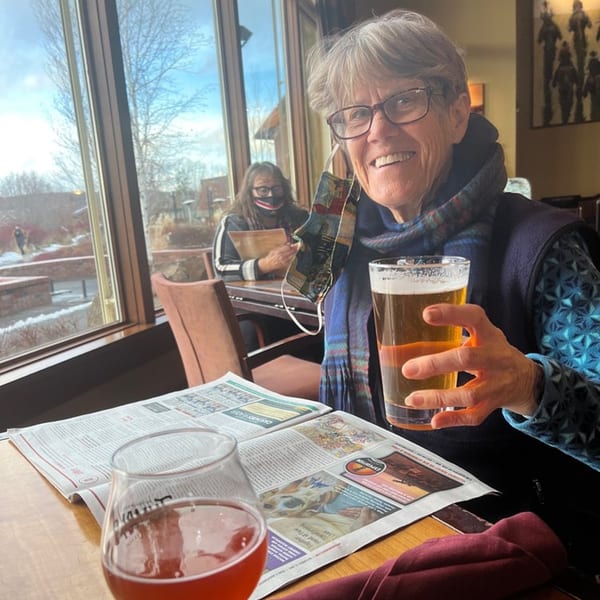 A smiling woman holding a beer at Greg's Grill, with a newspaper and another drink on the table.