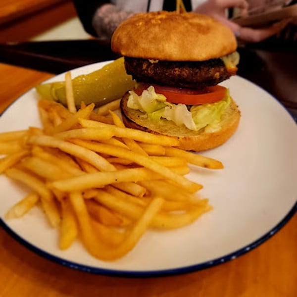 A Greg's Grill cheeseburger with lettuce, tomato, fries, and a pickle spear on a white plate.