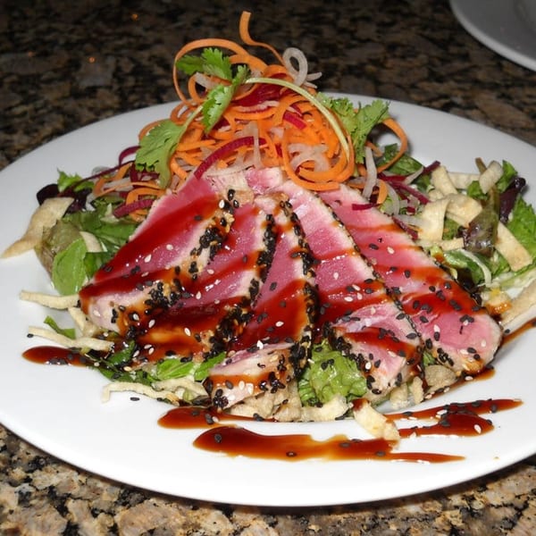 Seared tuna steak with sesame seeds, drizzled with sauce, on a bed of greens and shredded vegetables.