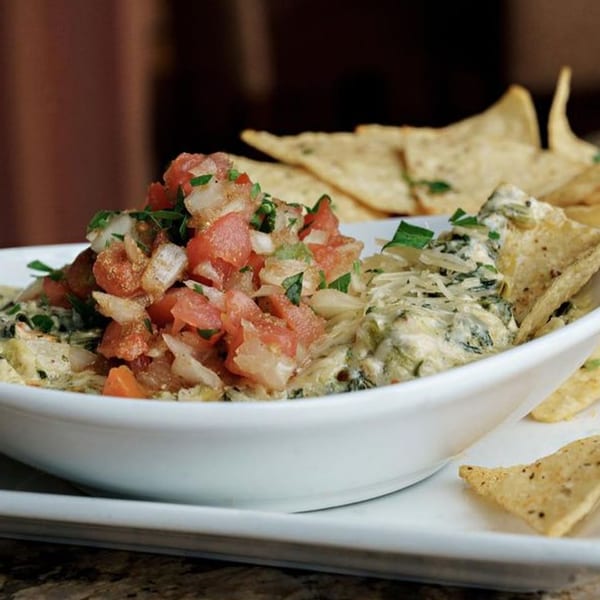 A white bowl of spinach dip topped with fresh salsa, surrounded by tortilla chips.