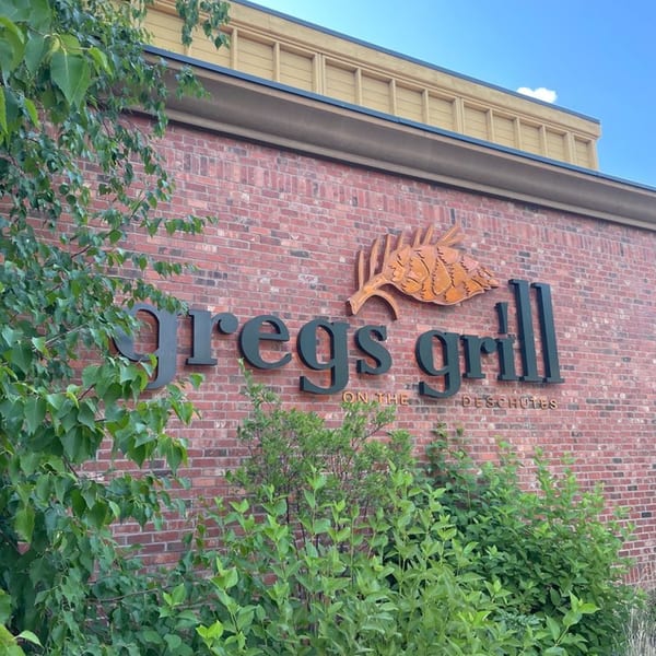 Greg's Grill sign on a brick building, partially obscured by green foliage.